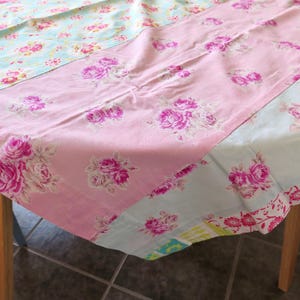 Peut inclure: Une nappe patchwork à motifs floraux. La nappe présente des sections de tissu rose, bleu clair et à motifs. La section rose présente de grandes roses roses. La nappe est drapée sur une table en bois.
