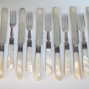 May include: A collection of antique silverware, including forks and knives. The forks have three tines and the knives have rounded blades. The handles are a pearlescent white color, and the metal is silver.