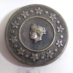 May include: A round, antique-style metal button with a central profile of a woman in a laurel wreath. The button is silver-toned with a dark background and a ring of star-shaped designs around the edge.