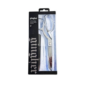 May include: A silver 8-inch Gingher knife-edge dressmaker shears in a clear plastic package with a black background. The package has the brand name "Gingher" printed in white.