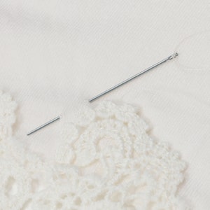 May include: A close-up of a white crochet lace trim with a silver needle lying on top of it.
