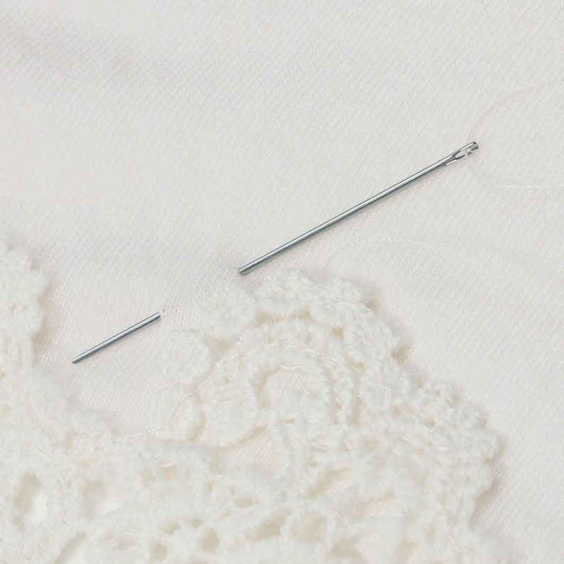 May include: A close-up of a white crochet lace trim with a silver needle lying on top of it.