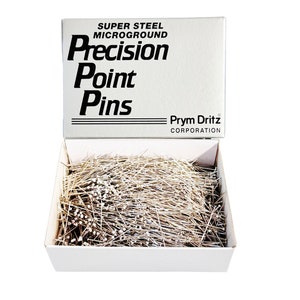 May include: A white box with the text "Super Steel Microground Precision Point Pins" and "Prym Dritz Corporation" on the lid. The box is open and filled with silver sewing pins.