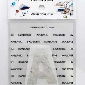 Puede incluir: Un paquete de plástico transparente que contiene una letra A blanca hecha de cristales brillantes. El paquete tiene el texto "SWAROVSKI" y "CREATE YOUR STYLE" impreso en él.