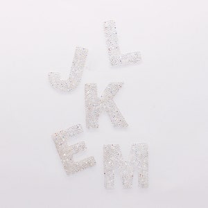 May include: White letters J, L, K, E, and M made of rhinestones. The letters are arranged in a random order.