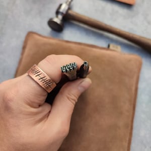 May include: A copper-colored ring with etched lines, held with metal tools and a hammer. The tools have textured ends. A brown leather pouch is in the background. The tools are likely for leatherworking or metal stamping.