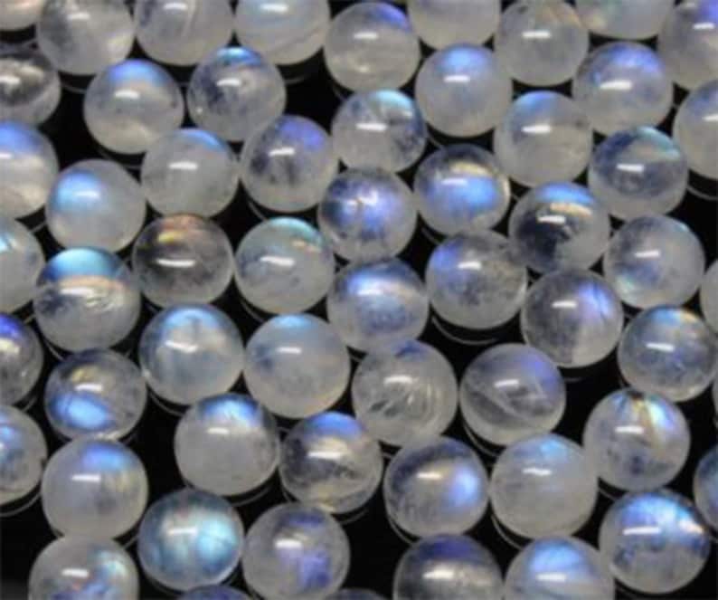 Puede incluir: Una colecci&oacute;n de cuentas redondas de piedra lunar iridiscente. Las cuentas son blancas con un brillo azul y est&aacute;n dispuestas en una vista de primer plano.