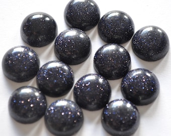 May include: A collection of twelve dark blue, polished cabochon gemstones with a sparkling, glittery appearance. The stones are round and domed, with a smooth, reflective surface. The gemstones are arranged on a white background.