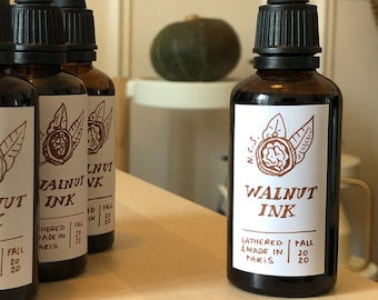 以下が含まれることがあります： 黒いスポイトが付いた茶色のガラス瓶のコレクションで、「Walnut Ink」とラベルされています。 各ラベルにはクルミのイラストと、インクがパリで2020年秋に収集および製造されたことを示すテキストが記載されています。 背景には小さな緑色のカボチャが見えます。