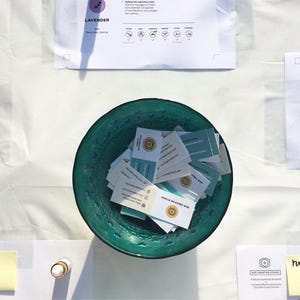 May include: A teal bowl filled with small white cards with gold lettering. The cards are stacked and scattered in the bowl. The bowl is on a white tablecloth with other papers and small bottles.