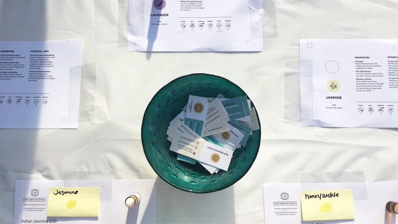 May include: A teal bowl filled with small white cards with gold lettering. The cards are stacked and scattered in the bowl. The bowl is on a white tablecloth with other papers and small bottles.