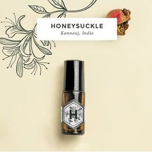 May include: A small brown glass bottle with a black rollerball top. The bottle has a white label with a black hexagonal design and the letter "H" inside. The label also says "Honeysuckle Kannauj, India".
