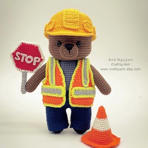 Pode incluir: Um urso de peluche de croch&ecirc; marrom vestindo um capacete amarelo, um colete de seguran&ccedil;a laranja e cal&ccedil;as azuis. O urso est&aacute; segurando uma placa de parada vermelha e est&aacute; de p&eacute; ao lado de um cone de tr&acirc;nsito laranja.