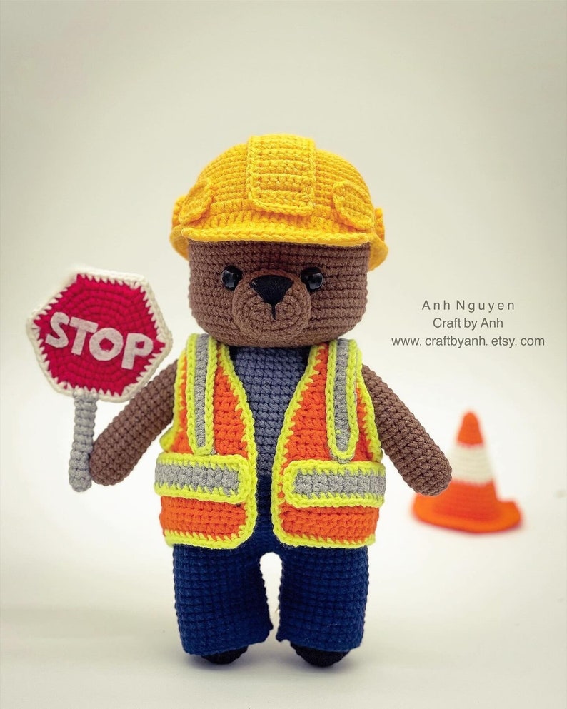 Pode incluir: Um urso de peluche de croch&ecirc; vestindo um capacete de constru&ccedil;&atilde;o amarelo, um colete de seguran&ccedil;a laranja e cal&ccedil;as jeans azuis. O urso est&aacute; segurando uma placa de parada vermelha. H&aacute; um cone de tr&acirc;nsito no fundo.
