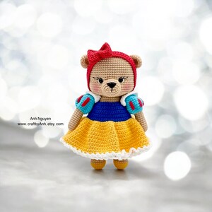 May include: A crocheted teddy bear dressed as Snow White. The bear is wearing a yellow dress with a blue bodice and a red bow in its hair. The bear is standing on a white background.