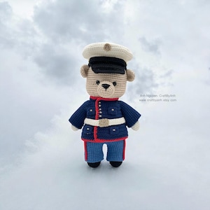 May include: A handmade crochet teddy bear wearing a navy blue uniform with red trim, a white belt, and a matching hat. The bear's uniform has gold buttons and is paired with blue pants. The bear is standing against a cloudy sky.
