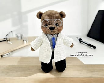May include: A brown crocheted teddy bear wearing a white doctor's coat, a blue tie, and round glasses. The bear is standing on a wooden surface.