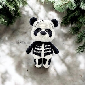May include: A crocheted panda bear wearing a skeleton costume. The panda is white with black eyes, nose, and ears. The skeleton costume is black with white bones. The panda is standing on a white surface.