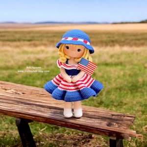 Op de afbeelding: Een gehaakte pop met een blauw-rood gestreept jurkje en een blauwe zonnehoed, die een kleine Amerikaanse vlag vasthoudt. De pop zit op een houten bankje in een veld.