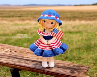 Può includere: Una bambola all'uncinetto con un vestito a righe blu e rosse e un cappello da sole blu, che tiene una piccola bandiera americana. La bambola è seduta su una panchina di legno in un campo.