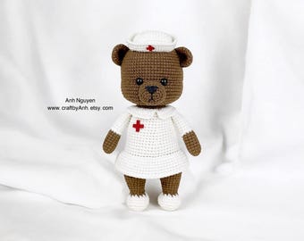 May include: A brown crocheted teddy bear dressed as a nurse. The bear is wearing a white dress with a red cross on the chest, a white hat with a red cross, and white shoes.