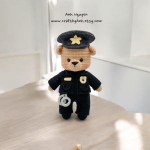 Puede incluir: Un oso de peluche de crochet vestido como policía. El oso lleva un uniforme negro con una insignia, un sombrero negro con una estrella dorada y esposas.