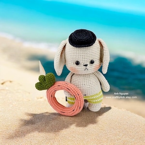May include: A crocheted bunny wearing a black hat and yellow and white striped swim trunks holds a pink and green inflatable ring. The bunny is on a sandy beach with blue water in the background.