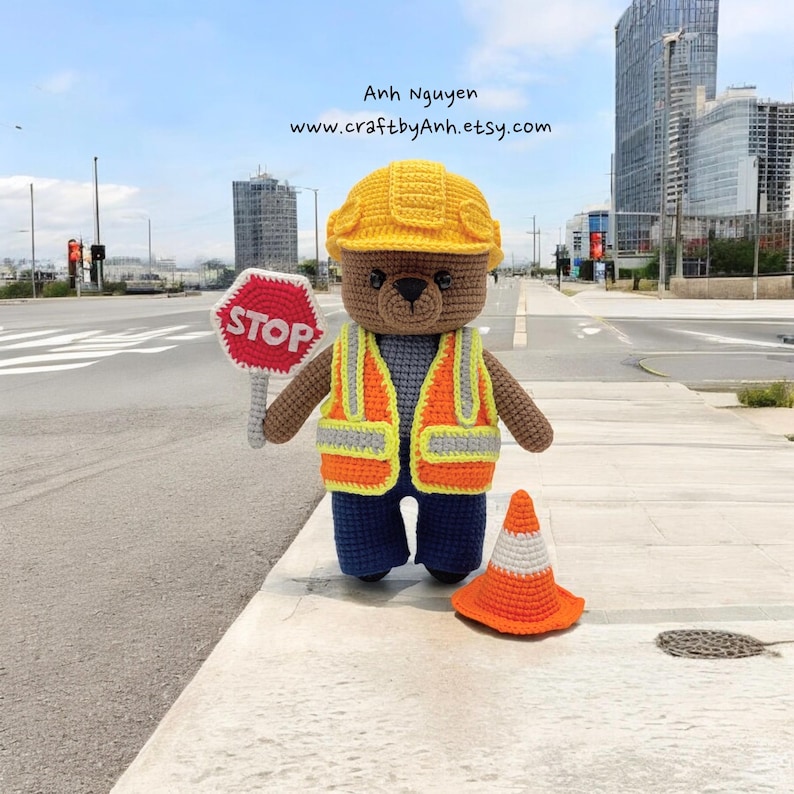 Pode incluir: Um urso de peluche de croch&ecirc; marrom vestindo um capacete amarelo, um colete de seguran&ccedil;a laranja e cal&ccedil;as azuis. O urso est&aacute; segurando uma placa de parada vermelha e est&aacute; de p&eacute; ao lado de um cone de tr&acirc;nsito laranja. O urso est&aacute; em uma cal&ccedil;ada em frente a uma rua da cidade.