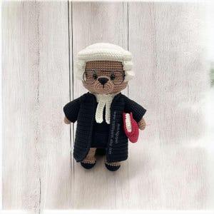 Pode incluir: Urso de pelúcia de crochê vestido de juiz, usando uma toga preta, peruca branca e óculos redondos. O urso segura um livro vermelho. O fundo é um padrão de grão de madeira clara.
