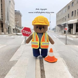 Pode incluir: Um urso de peluche de croch&ecirc; marrom vestindo um capacete amarelo, um colete de seguran&ccedil;a cinza e laranja e jeans azul. O urso est&aacute; segurando uma placa de parada vermelha e est&aacute; de p&eacute; ao lado de um cone de tr&acirc;nsito laranja. O urso est&aacute; em uma faixa de pedestres em frente a uma rua da cidade.