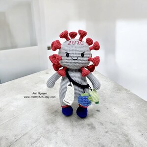 May include: A gray crocheted plush toy shaped like a coronavirus with red spikes. The toy has a face with a frown and the number 2020 on its head. It is wearing a blue mask and carrying a small blue globe.