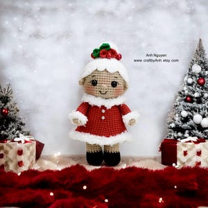 Puede incluir: Una muñeca de elfo de Navidad de ganchillo que lleva un vestido rojo con ribetes blancos, un sombrero blanco con acebo verde y zapatos negros. La muñeca está de pie sobre una alfombra de piel roja frente a dos árboles de Navidad en miniatura.