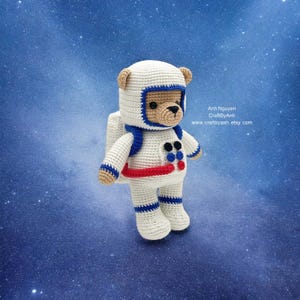 Puede incluir: Un oso de peluche tejido a crochet vestido de astronauta, con un traje espacial blanco con detalles azules y un cinturón rojo. El oso está de pie sobre un fondo azul con estrellas blancas.