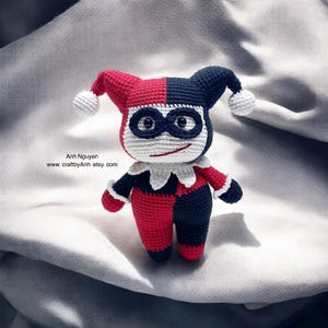 May include: A crocheted stuffed toy of Harley Quinn, a character from the DC Comics universe. The toy is wearing a red and black jester costume with a white collar and a black mask with white eyes. The toy has a white and black jester hat with a white pom-pom on top.