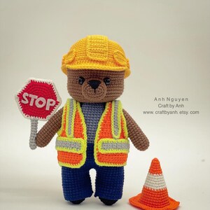 Pode incluir: Um urso de peluche de croch&ecirc; marrom vestindo um capacete de constru&ccedil;&atilde;o amarelo, um colete de seguran&ccedil;a laranja e jeans azul. O urso est&aacute; segurando uma placa de parada vermelha e est&aacute; de p&eacute; ao lado de um cone de tr&acirc;nsito laranja.