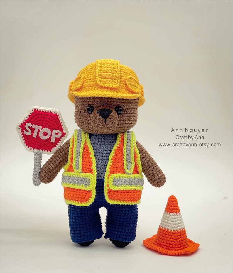Pode incluir: Um urso de peluche de croch&ecirc; marrom vestindo um capacete de constru&ccedil;&atilde;o amarelo, um colete de seguran&ccedil;a laranja e jeans azul. O urso est&aacute; segurando uma placa de parada vermelha e est&aacute; de p&eacute; ao lado de um cone de tr&acirc;nsito laranja.