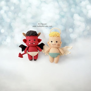 Puede incluir: Un diablo rojo de crochet y un ángel beige de crochet con un halo amarillo, ambos con pañales. El diablo tiene alas negras y una cola, mientras que el ángel tiene alas blancas. El fondo es azul claro con bokeh blanco.