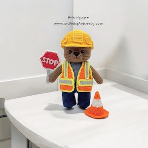 May include: A crocheted teddy bear dressed as a construction worker. The bear is wearing a yellow hard hat, an orange safety vest, blue trousers, and holding a red stop sign. There is an orange traffic cone on the table.