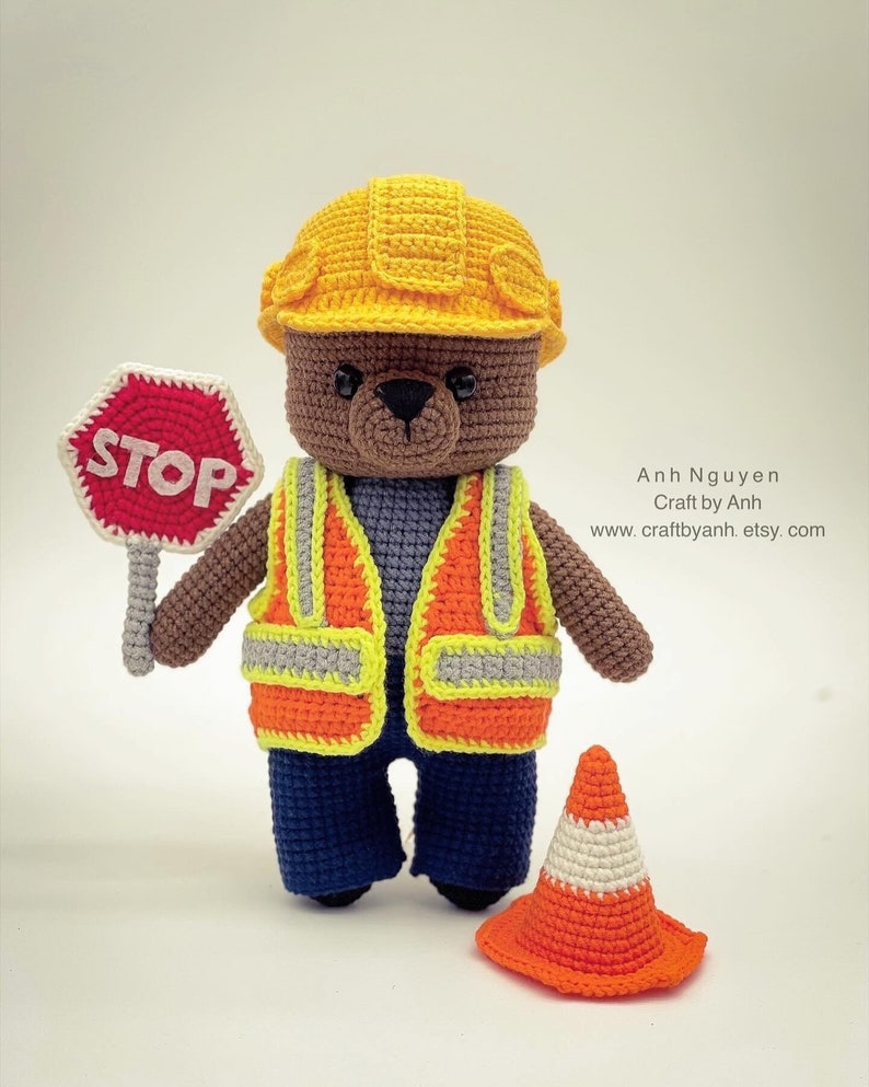 Pode incluir: Um urso de peluche de croch&ecirc; marrom vestindo um capacete amarelo, um colete de seguran&ccedil;a laranja e cal&ccedil;as azuis. O urso est&aacute; segurando uma placa de parada vermelha e est&aacute; de p&eacute; ao lado de um cone de tr&acirc;nsito laranja.