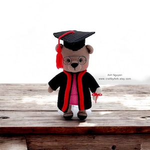 Puede incluir: Un oso de peluche de crochet marrón que lleva una gorra y una bata de graduación negras, gafas rojas y sostiene un diploma rojo. El oso está de pie sobre una superficie de madera.