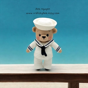 May include: A crocheted teddy bear wearing a white sailor hat, white sailor uniform with blue and white stripes, and a black tie. The bear is standing on a wooden surface.