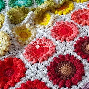 May include: A crocheted blanket featuring a white background and colourful flower motifs. The flowers are in shades of red, orange, yellow, green, and blue.