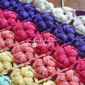 May include: A close-up of a crocheted blanket with a repeating flower pattern in shades of pink, purple, white, and yellow. The blanket is made with a textured stitch that creates a 3D effect.
