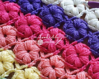 May include: A close-up of a crocheted blanket with a repeating flower pattern in shades of pink, purple, white, and yellow. The blanket is made with a textured stitch that creates a 3D effect.