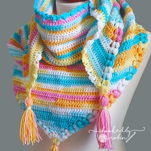 May include: A vibrant, hand-crocheted shawl in yellow, pink, blue, and white. The shawl has a striped design, decorative edging, and tassels. The text "hooked by robin" is visible. The shawl is a triangular shape.