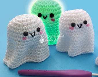 May include: Three crocheted ghost amigurumi toys. Two are white with black eyes and pink cheeks. One is green with black eyes and pink cheeks. The green ghost is glowing. The text "hooked by robin" is visible in the image.