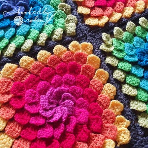 May include: A close-up of a colorful crochet blanket with a rainbow pattern. The blanket is made of multiple crocheted flowers in different colors, including red, orange, yellow, green, blue, and purple.