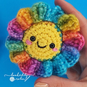 May include: A crocheted rainbow flower with a yellow smiling face in the centre. The flower has a blue, green, pink, orange, and yellow colour scheme.