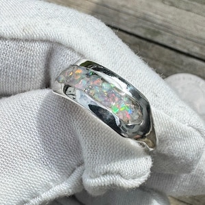 May include: A silver ring with an elongated, curved design. The ring features a central inlay of iridescent, multicolored opal. The opal displays flashes of green, blue, and orange. The ring is set against a white background.