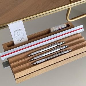 May include: A wooden desk organiser with a ruler, pencils, and eraser. The organiser has a slot for a notepad with the text "MELDE MADE" and a logo. The ruler is marked with red and white lines. The pencils are silver.
